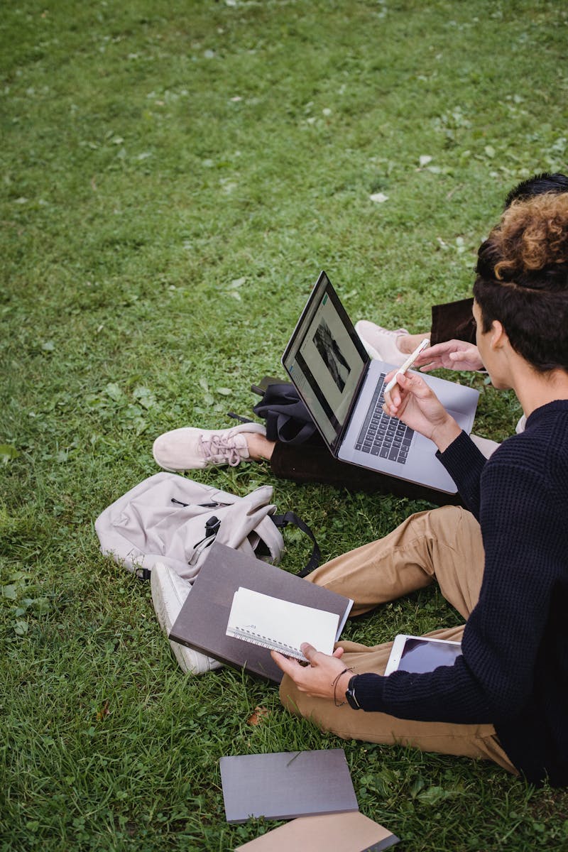 Két diák tanul együtt egy laptopon a szabadban, könyvekkel körülvéve.